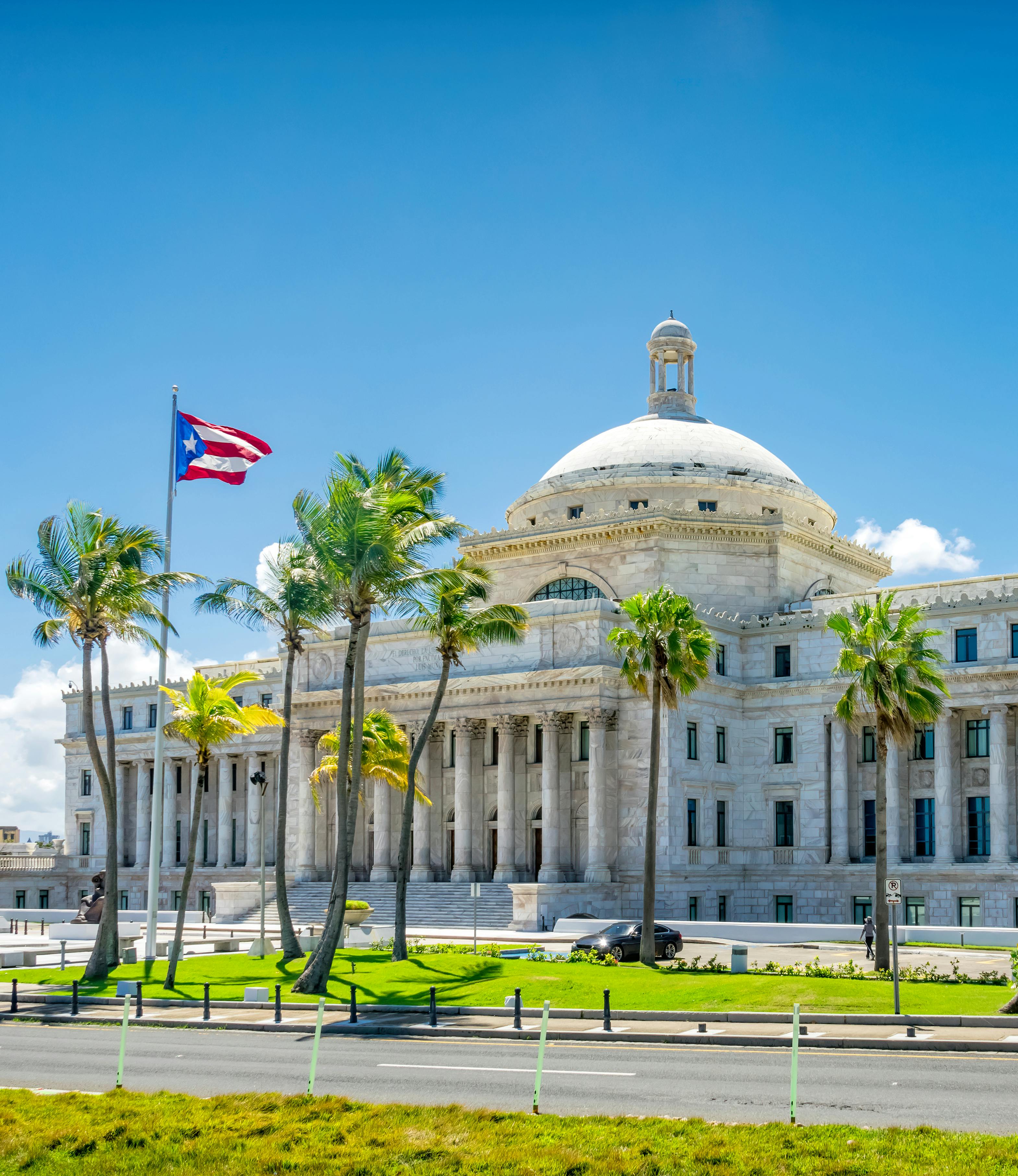 The front of the Puerto Rico Capitol building in San Juan on a sunny day, with columns in the front and a dome; a Puerto Rican flag flies from a flagpole in front of the building; palm trees line the street from side to side.