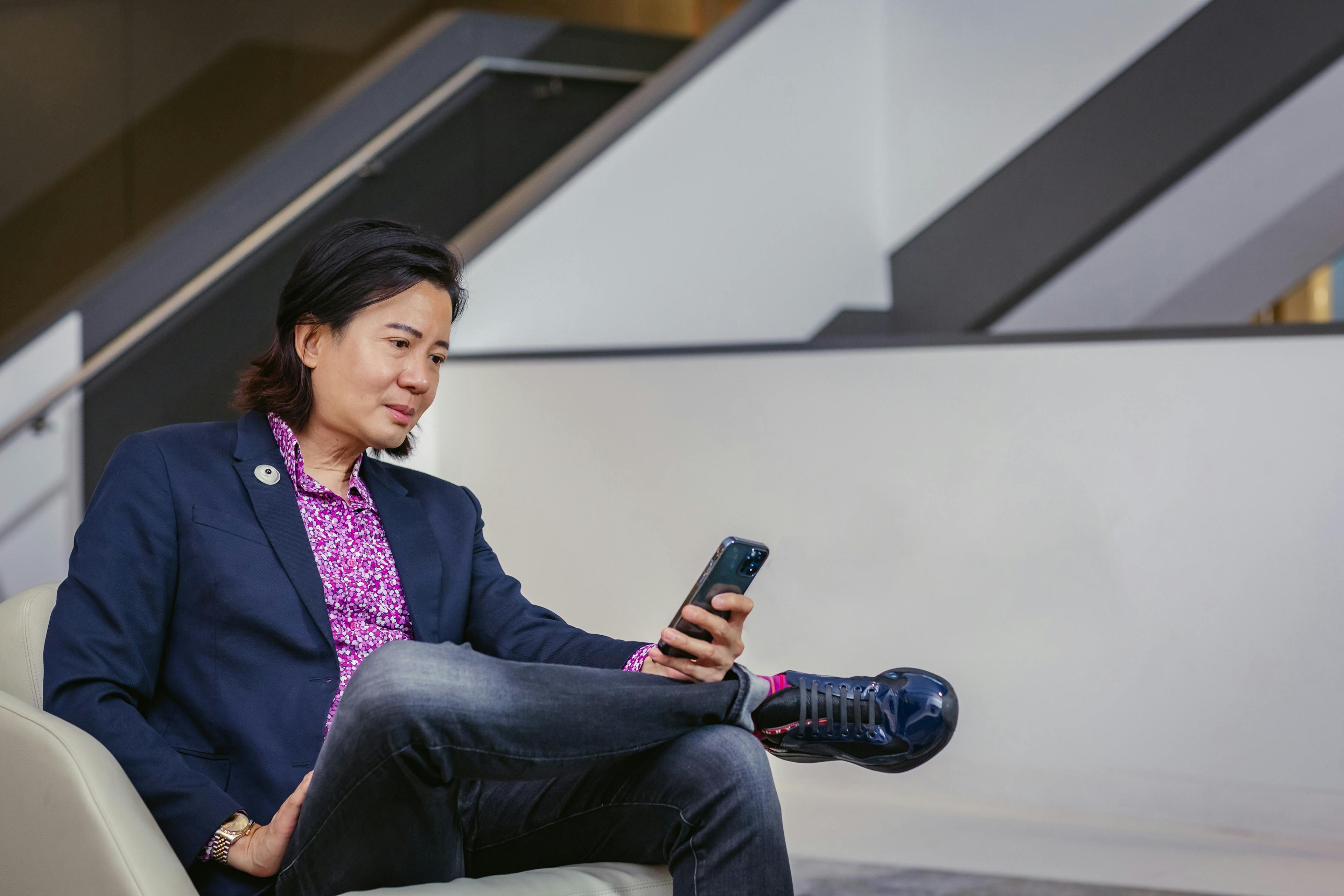A person in a purple shirt and blue blazer sitting on a couch and looking at their phone.
