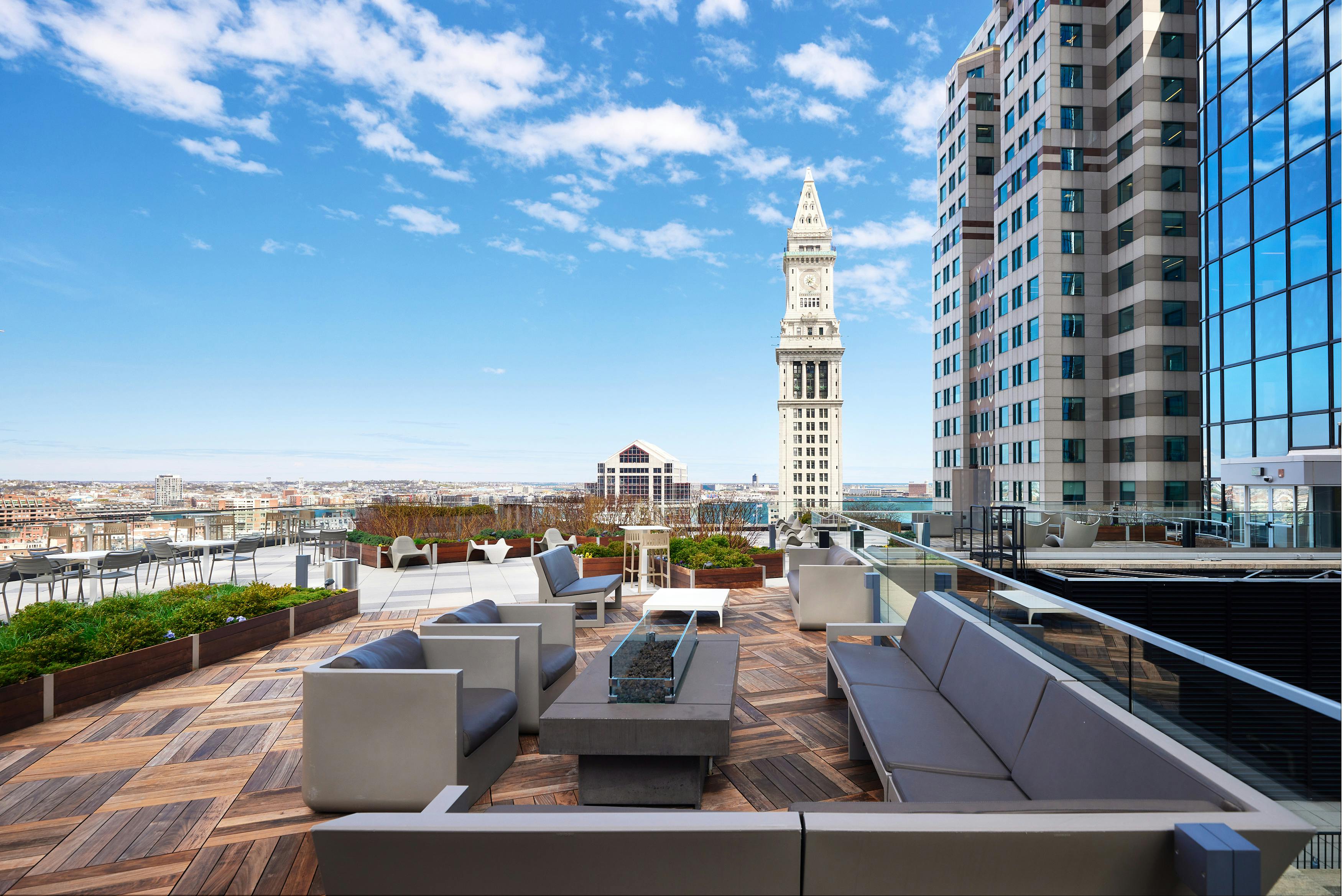 Nixon Peabody's Boston office rooftop lounge; couches and single chairs surround a long firepit/coffee table; other seating appears in the background; beyond the rooftop, the Boston Custom House clock tower, with the city stretching away into the distance, blue skies above