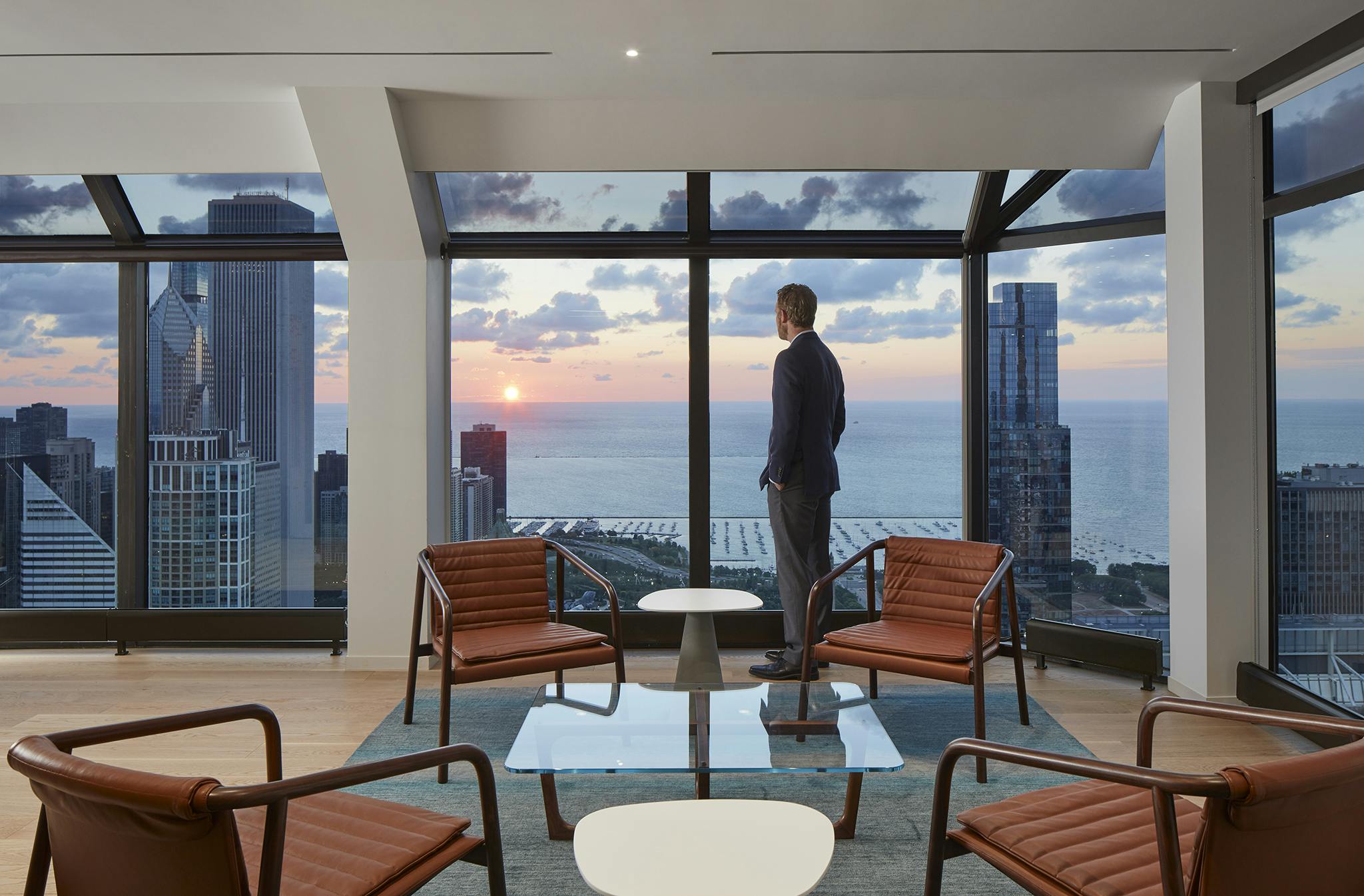 A seating area in Nixon Peabody's Chicago office, with a square glass coffee table surrounded by four brown leather chairs; a person stands behind them, looking through a panel of floor-to-ceiling windows, showing other Chicago skyscrapers and the sun setting over Lake Michigan