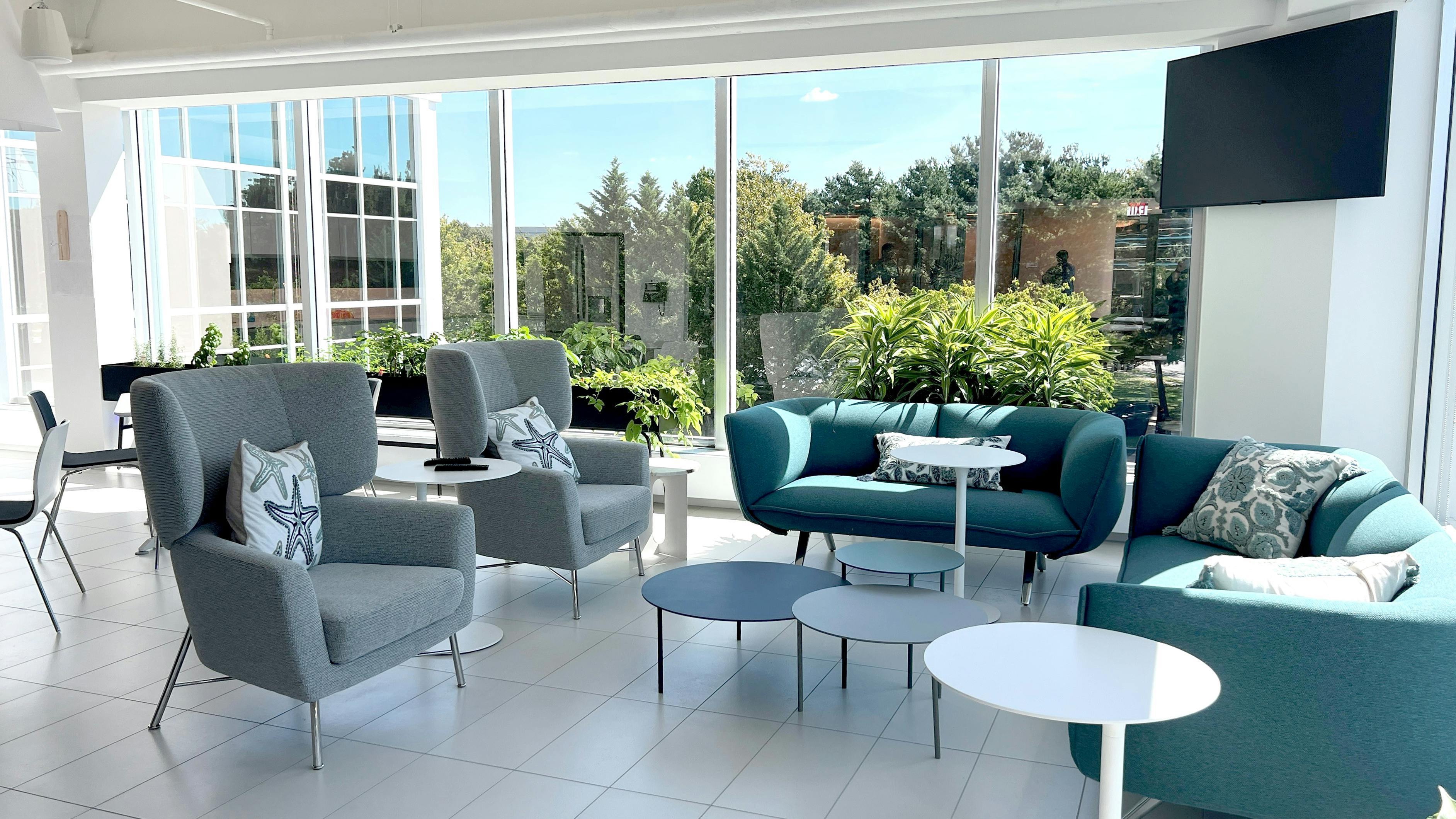 Lounge area with wingback chairs, a couch and loveseat, and small round side tables; floor-to-ceiling windows in the background show trees outside the building.