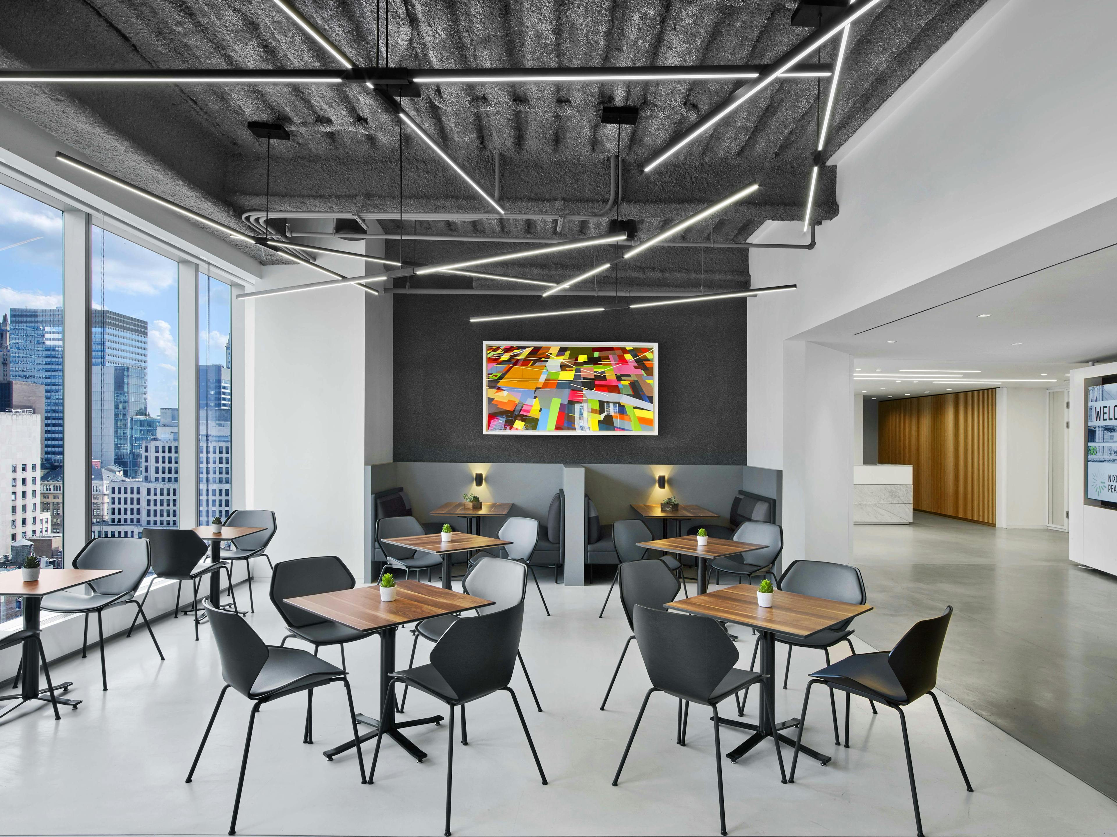 The cafe in Nixon Peabody's New York City office; two tables, each with four chairs, stand in the foreground; behind these are two booths, with a painting hanging on the wall behind; on left to the side are picture windows and small tables with chairs, and on the right can be seen the hallway leading to the reception desk