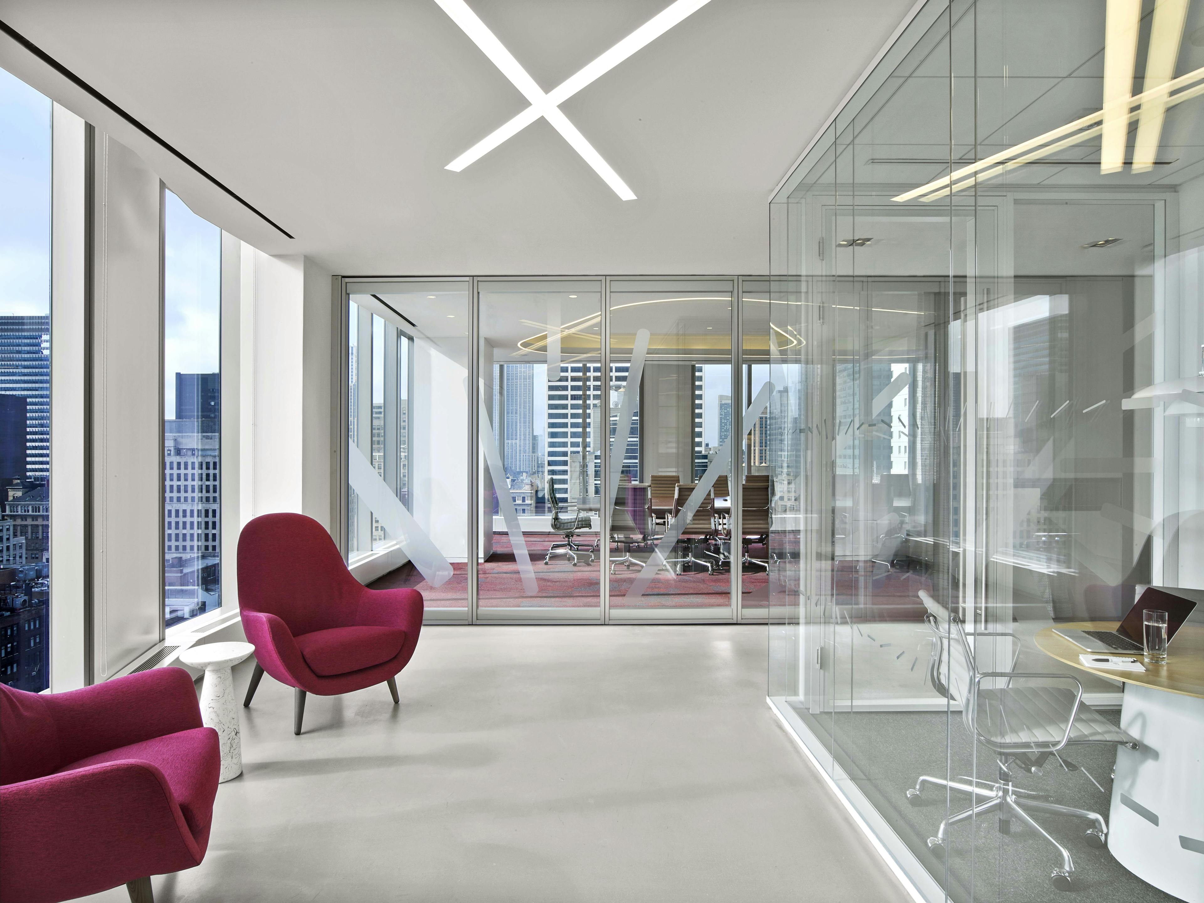 Seating area in Nixon Peabody's New York City office; two fuchsia easy chairs sit alongside picture windows to the left, a small occasional table between them; directly behind is an empty conference room with glass walls and an oblong table surrounded by chairs; on the right can be seen part of another, smaller, glass-enclosed conference room