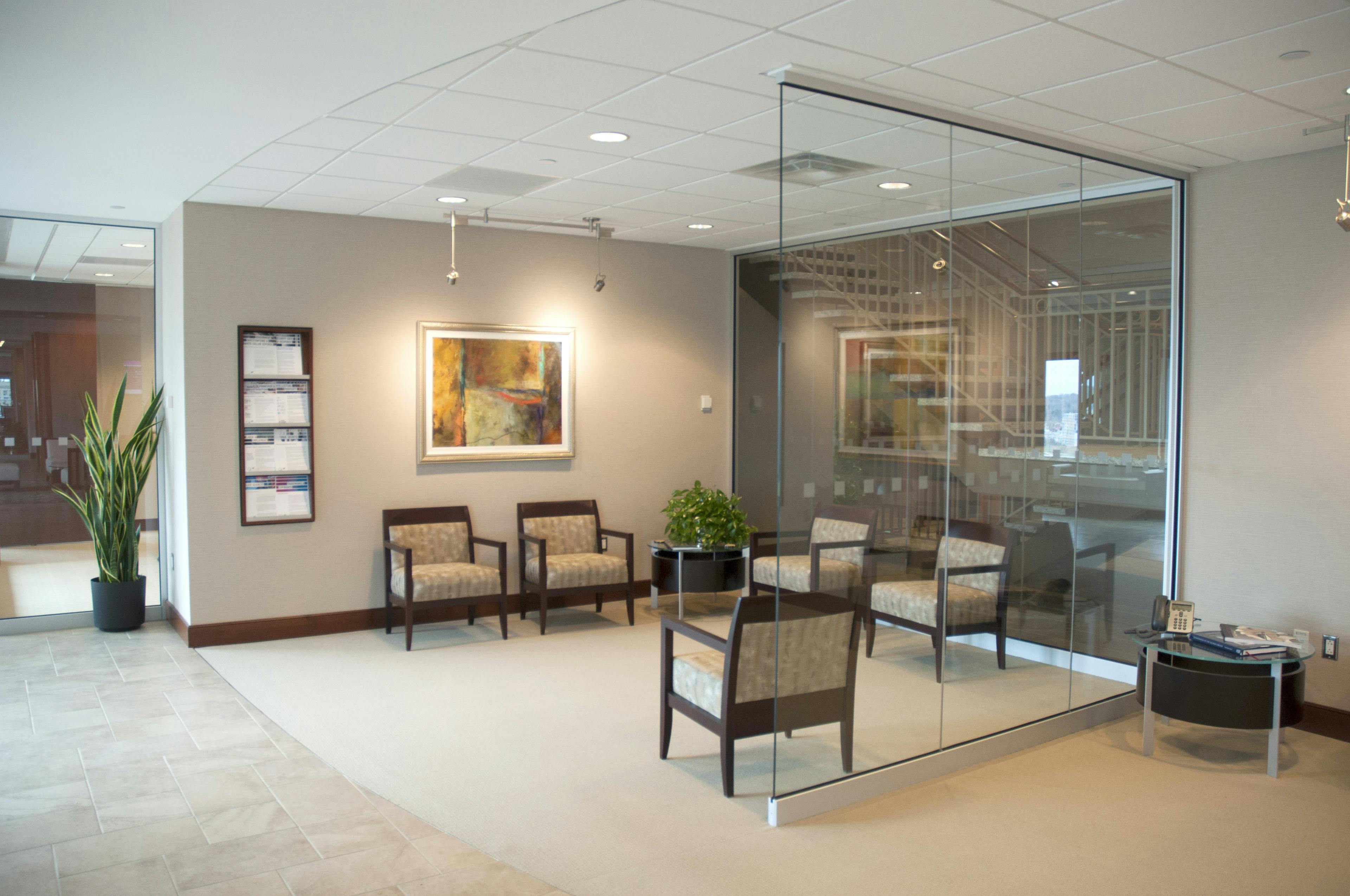 Reception seating area in Nixon Peabody's Rochester office; a small space, enclosed on three sides by two glass panels and one wall, contains five chairs and a small table on which is a plant; through one of the glass walls can be seen the office stairwell; a painting hangs on the wall, along with a rack of firm brochures
