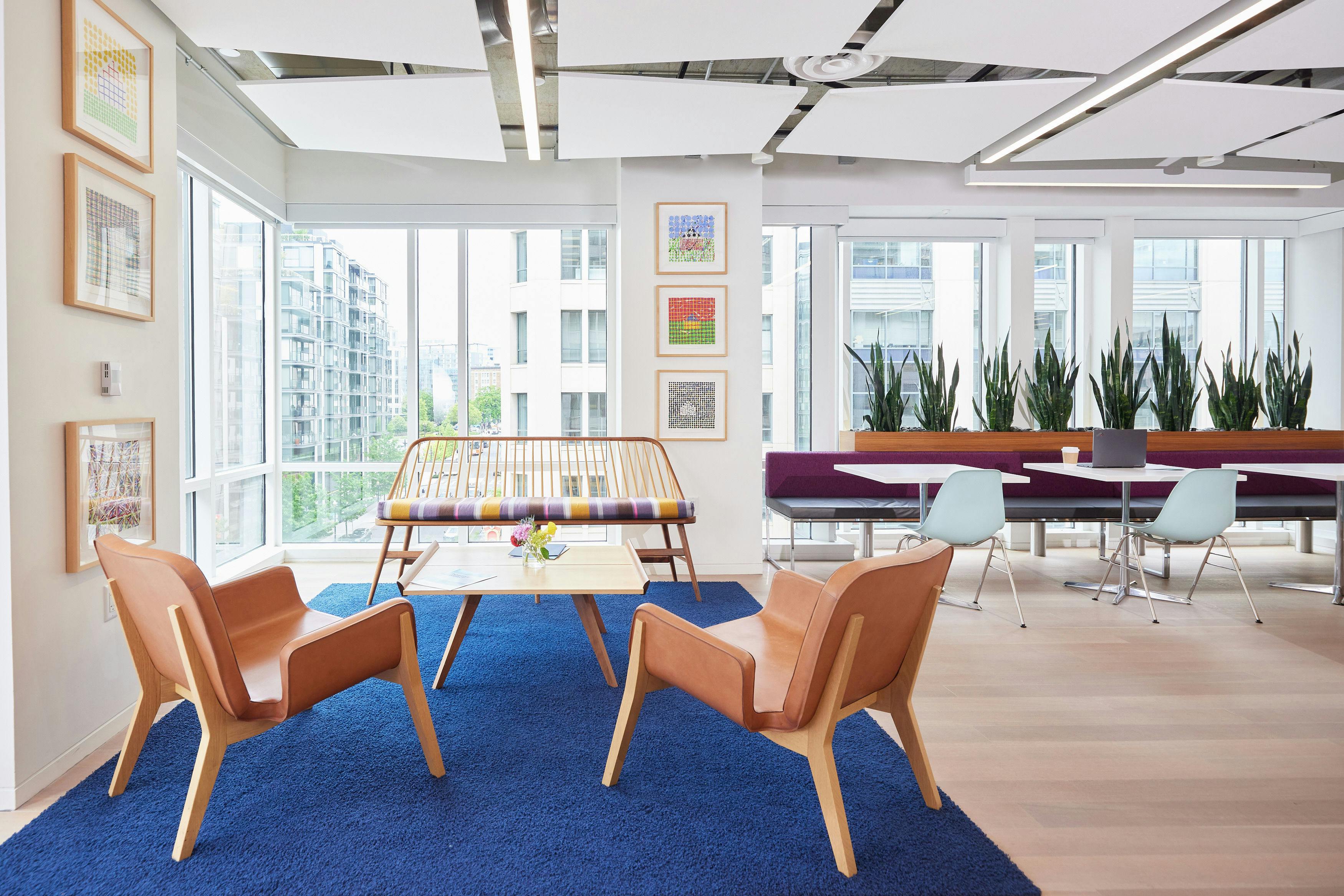 Corner of the lounge area in Nixon Peabody's DC office; on a royal blue area rug sits a table with two chairs with their backs to the viewer; facing the viewer on the other side of the table is a bench with a multi-colored cushion; additional cafe seating is visible in the back stretching to the right out of view; artwork adorns the walls and nearby buildings appear through the windows