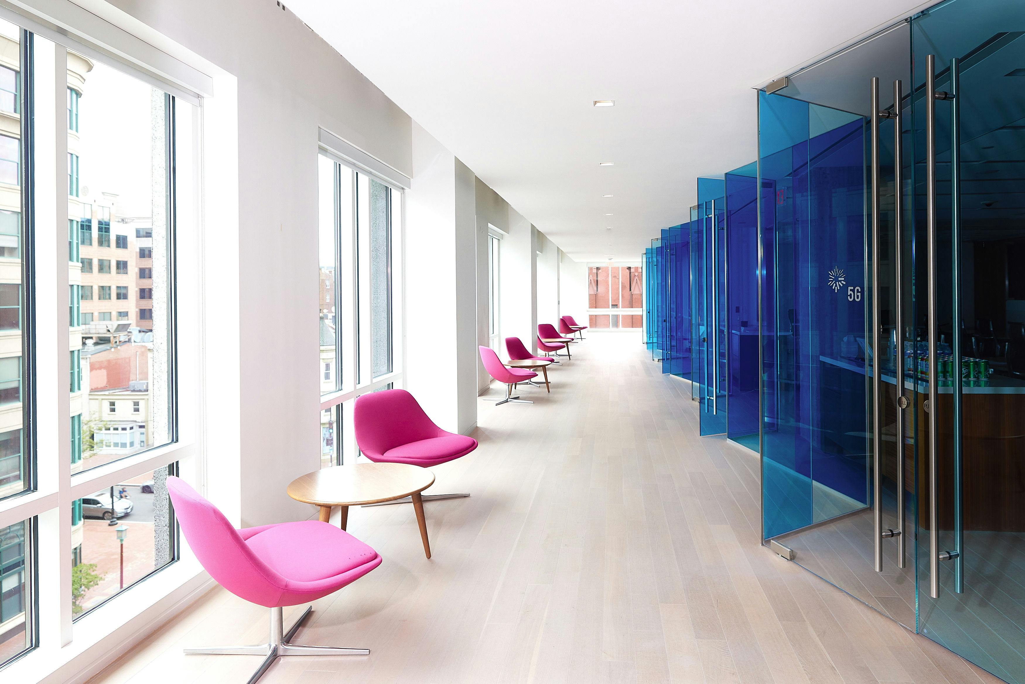 Hallway in Nixon Peabody's DC office; on the right are a series of small conference rooms, each one enclosed in blue-tinted glass; on the left pink chairs and small round wooden tables stretch into the distance along a row of floor-to-ceiling windows