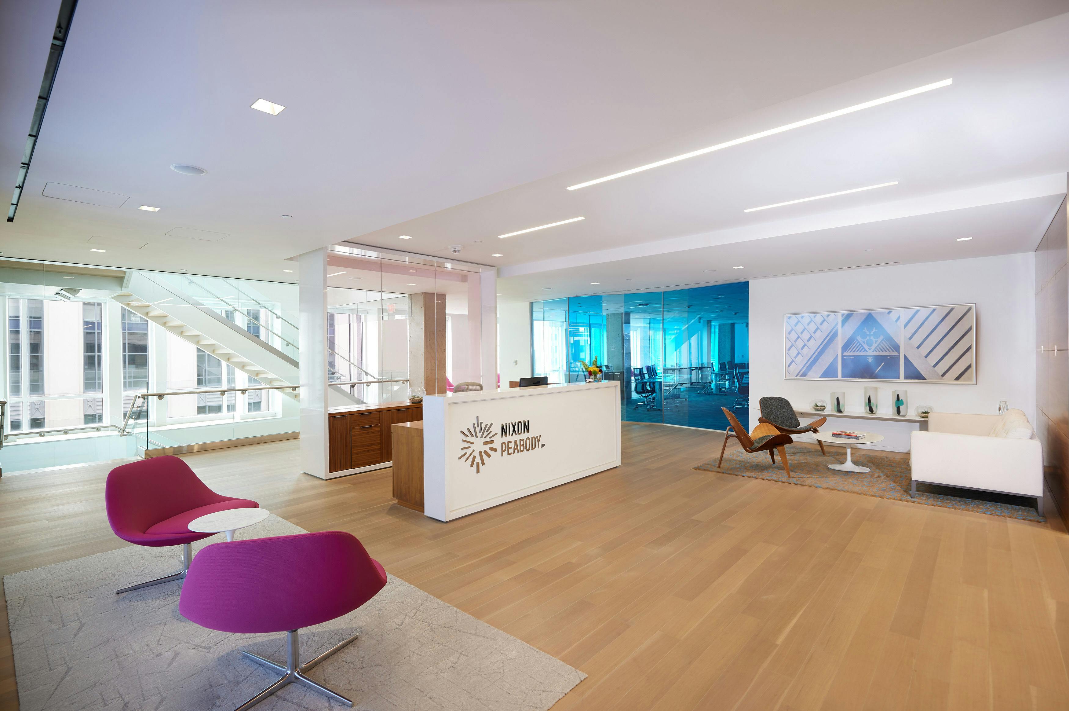 The reception area of Nixon Peabody's DC office; on the right in the foreground are two pink chairs with a small occasional table; in the middle ground is the white reception desk with the Nixon Peabody logo on the front; behind the desk is a glass-enclosed stairwell; in the background is another small seating area with a couch and two chairs, and a large conference room behind panels of blue-tinted glass