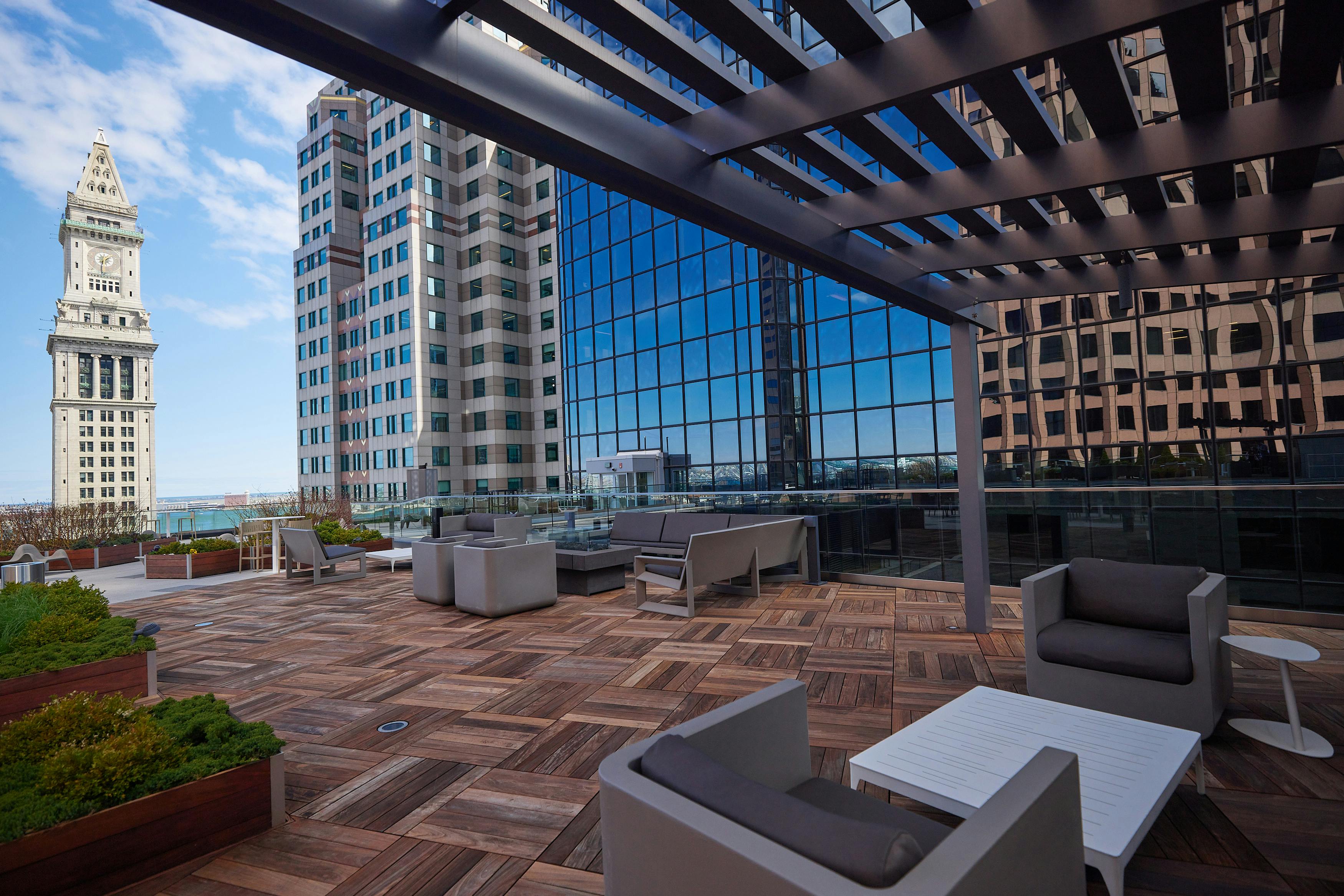 Nixon Peabody's Boston office rooftop lounge area, with wood-paneled floors, couches and easy chairs, and open-beam canopy, and long firepit coffee tables; the Boston Custom House Tower appears nearby beyond the edge of the rooftop
