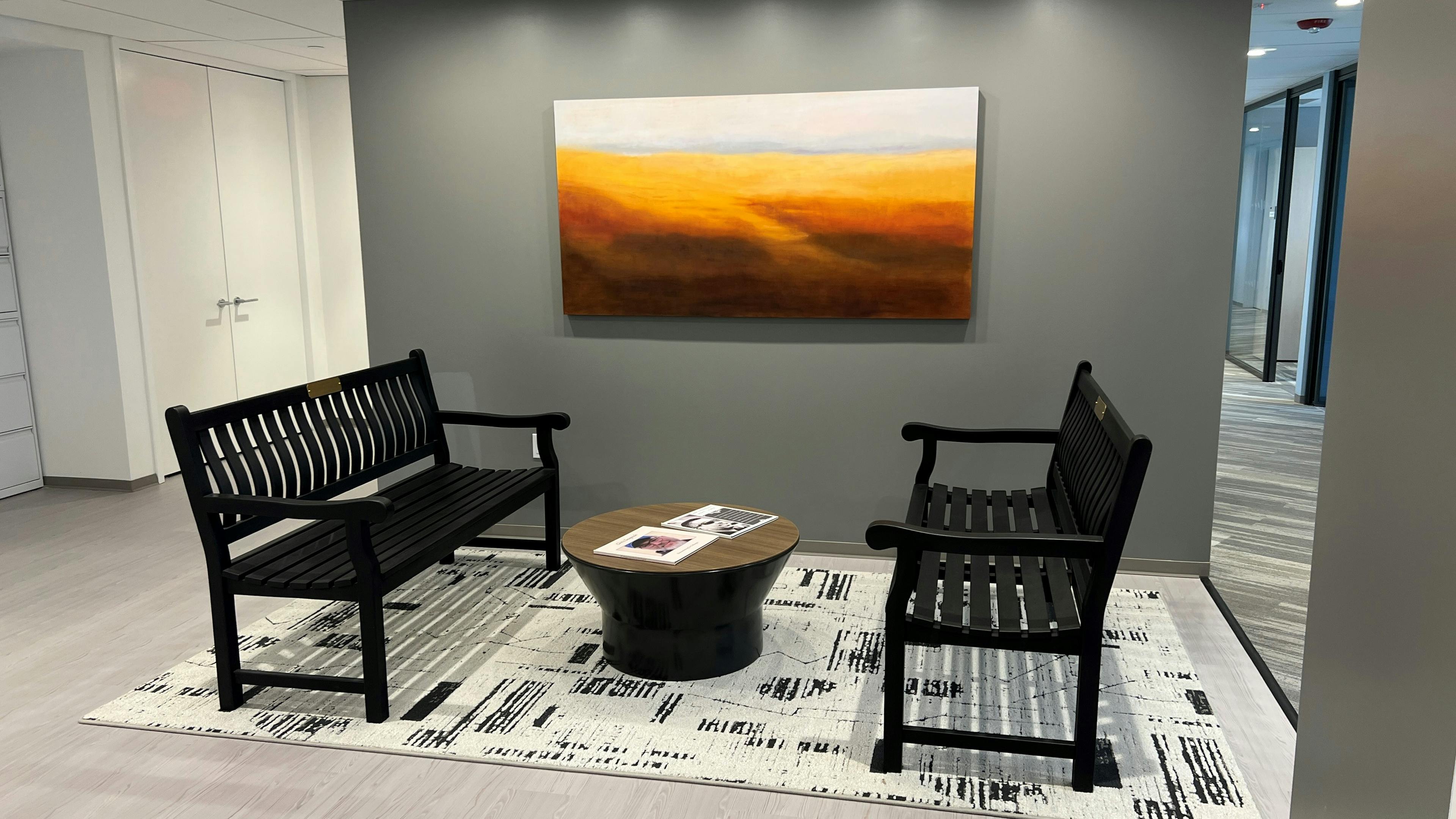 Two benches in an office waiting room area, a small round coffee table in between and a landscape painting on the wall facing the viewer