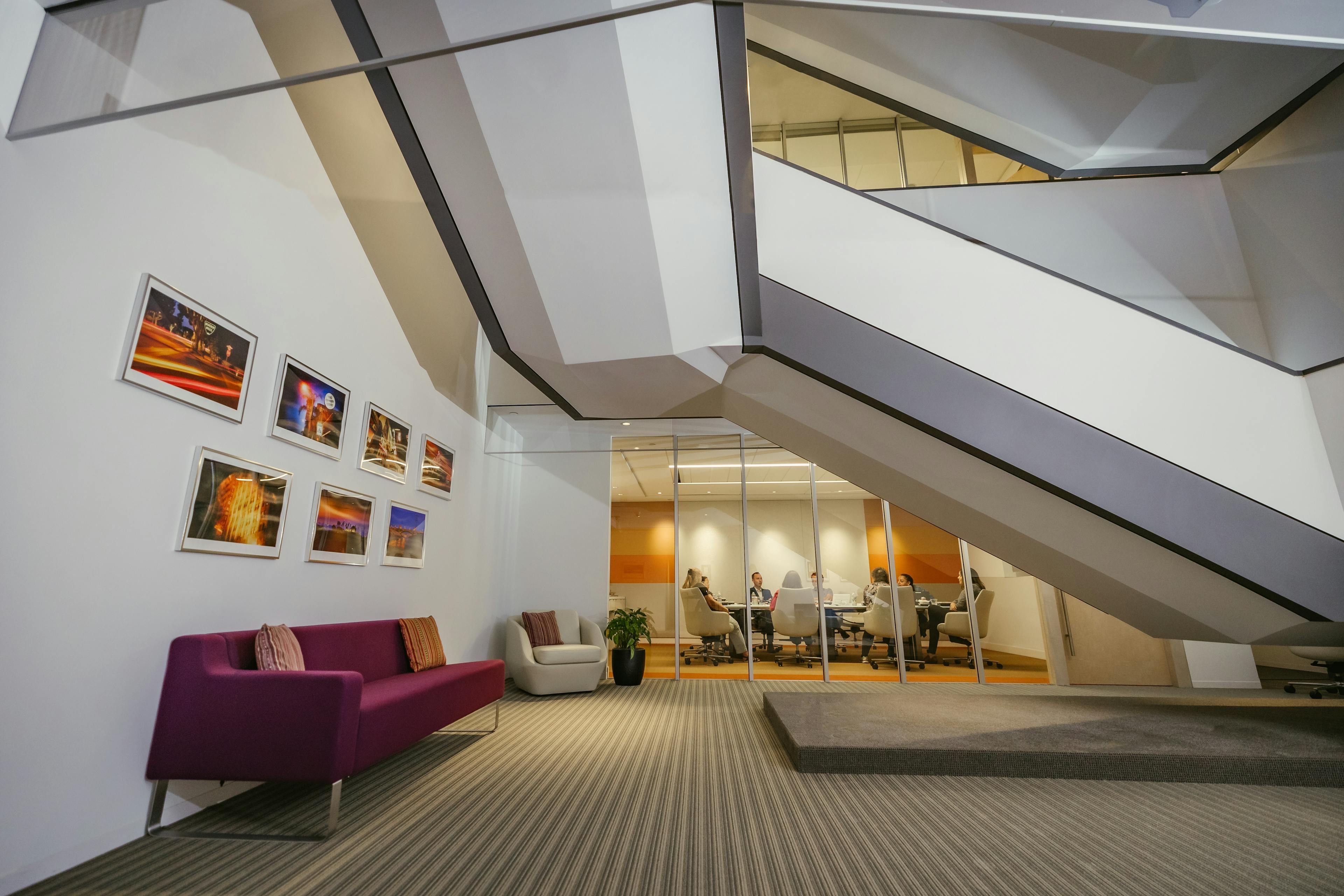 Interior of Nixon Peabody's Los Angeles office; on the left wall is a purple couch, behind which hang 7 framed photographs in two rows; a staircase leads up from right to left; in the background, looking under the staircase suspended above, a conference room can be seen through glass panels with a group of people sitting around an oblong table