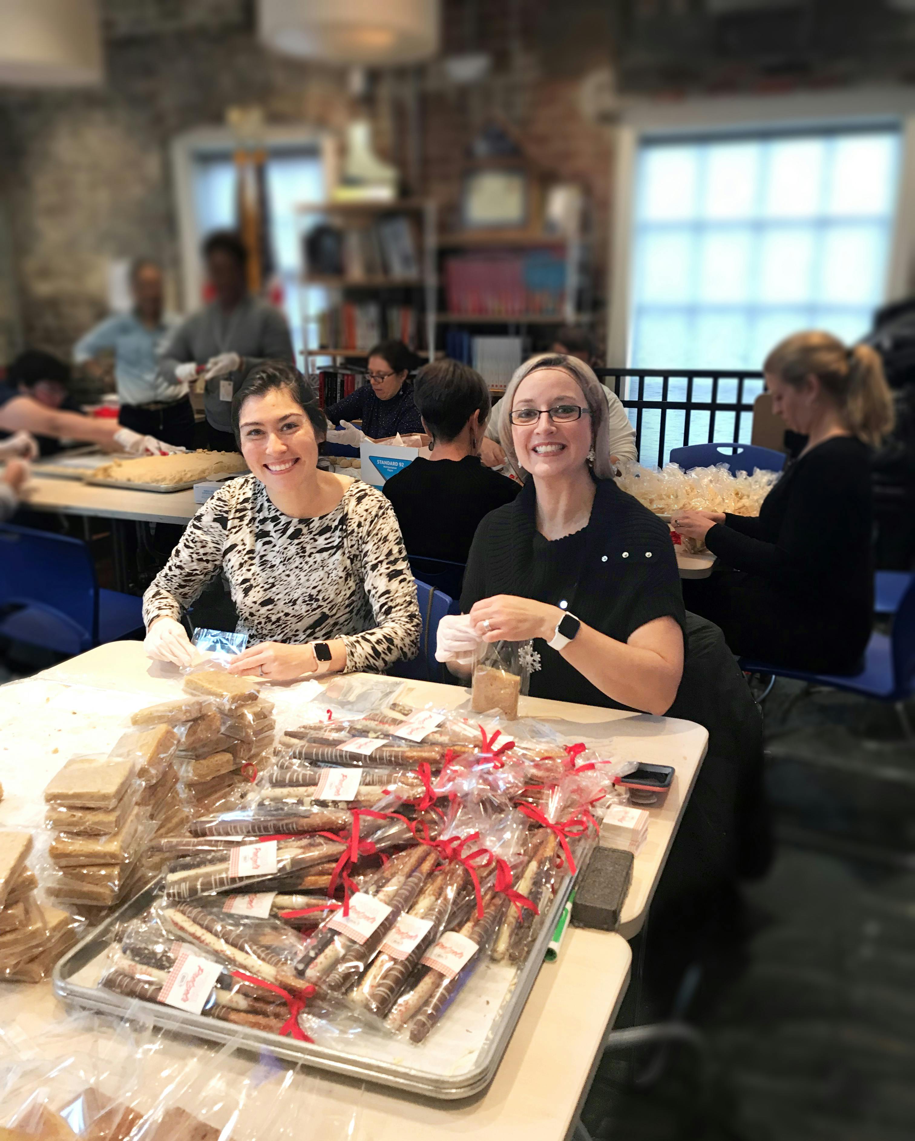 Two people smiling at the camera, seated at a table where they are packing cookies into plastic bags and tying them with a red ribbon. A pile of completed packages is on the table in front of them. In the background are other people engaged in similar activities.